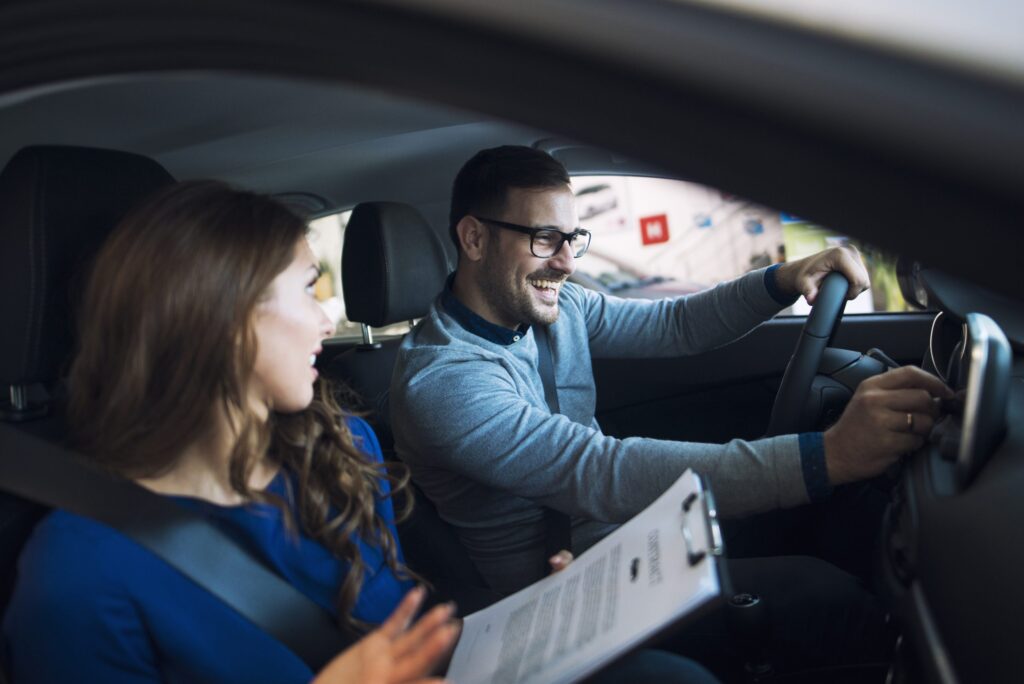 Homem e mulher dentro de um carro, com o homem ao volante sorrindo e ajustando o painel, enquanto a mulher observa, segurando uma prancheta com documentos. A imagem transmite um ambiente de aprendizado ou instrução sobre direção.