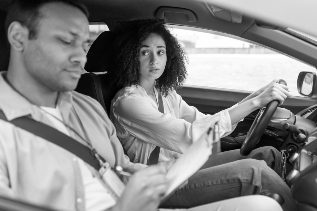 Cenário de aula de direção, com uma mulher ao volante e um homem ao lado, analisando um documento. Ambos parecem concentrados, refletindo a dinâmica de aprendizado e instrução em um carro.