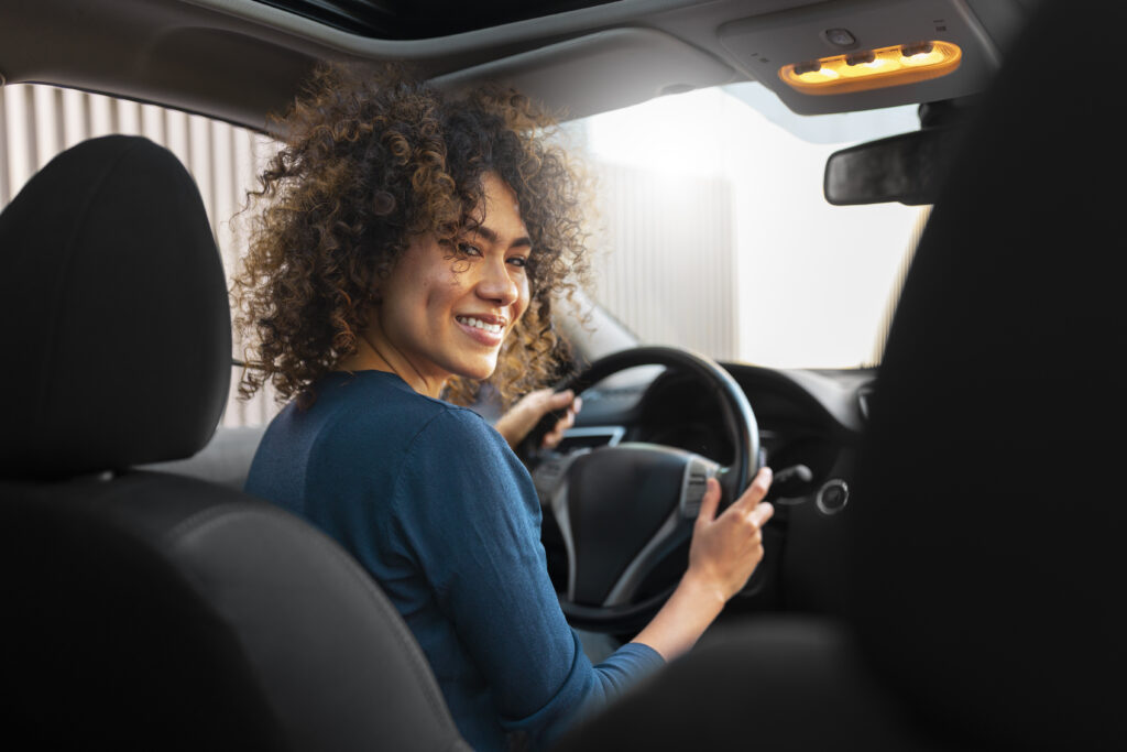 Mulher sorridente dirigindo um carro, vista do banco traseiro, com cabelo cacheado e usando uma blusa azul.